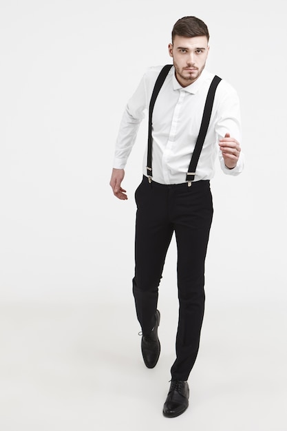 Full length vertical picture of concentrated attractive young unshaven male in elegant suit running against white studio wall background, his look expressing determination, seriousness and confidence
