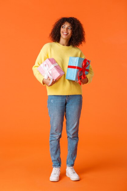 Полнометражный вертикальный выстрел мечтательной и симпатичной привлекательной афро-американской женщины, оглядывающейся вокруг, держа подарки, оранжевую стену.