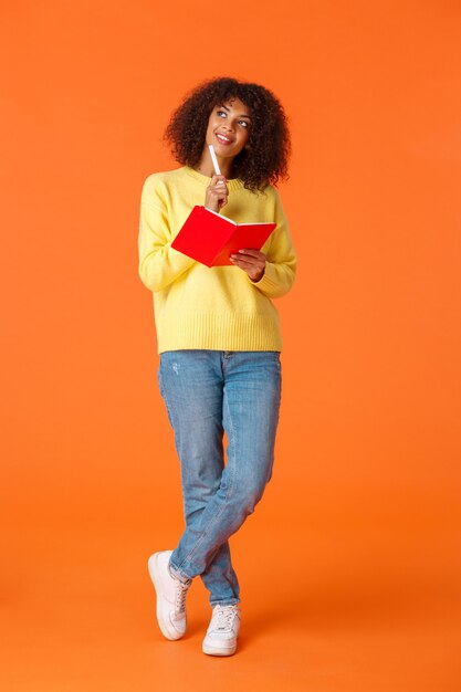 Full-length vertical shot dreamy and romantic cute girl making schedule, taking notes or to-do list, imaging something as writing in red cute notebook, touch chin with pen looking up thoughtful.