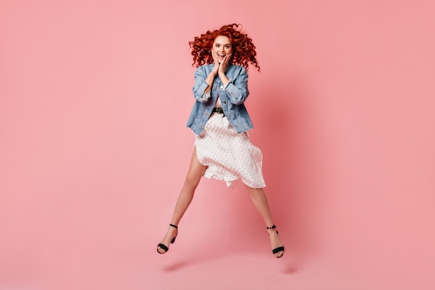 Free photo full length view of amazed ginger lady jumping on pink background. studio shot of active curly girl in denim jacket and high-heeled shoes.