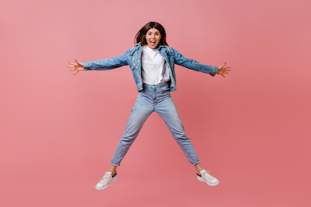 Full length view of amazing girl in denim attire waving hands on pink background. Young woman jumping and smiling.