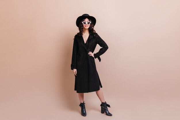 Full length view of asian woman in black coat. Studio shot of confident korean woman standing on beige background.