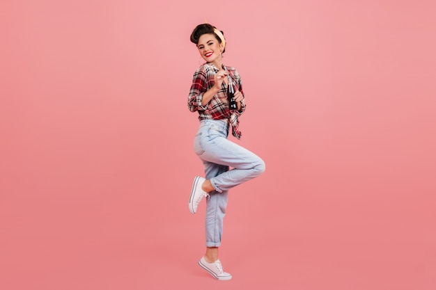 Full length view of blissful woman dancing on pink background. Studio shot of pinup lady in checkered shirt.