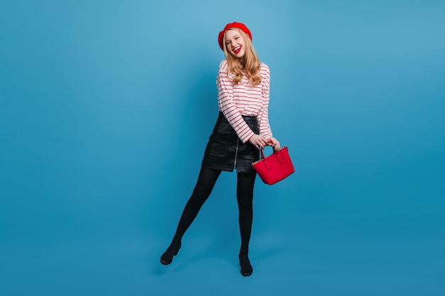 Full length view of carefree girl in beret and short skirt Studio shot of pretty blonde woman with red handbag