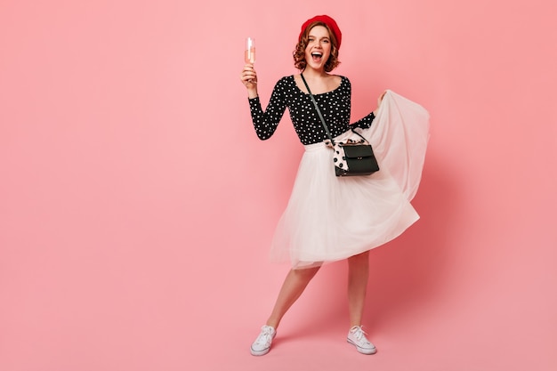 Full length view of cheerful french girl playing with skirt. Blissful curly woman in beret holding wineglass on pink background.