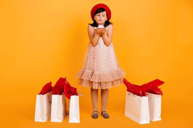 Free photo full length view of child in dress celebrating birthday studio shot of kid in beret blows out candle on cake