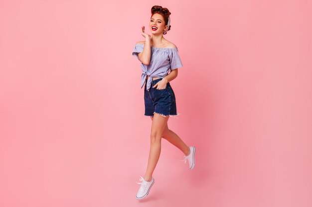 Full length view of cute pinup girl jumping on pink background Studio shot of glamorous ginger woman in denim shorts