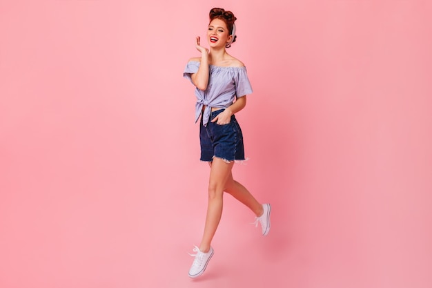 Full length view of cute pinup girl jumping on pink background Studio shot of glamorous ginger woman in denim shorts