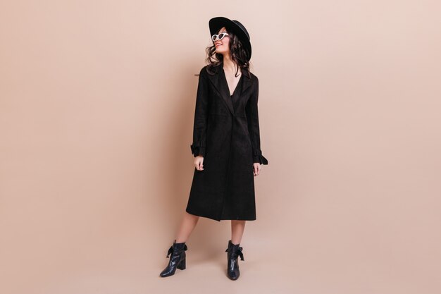 Full length view of dreamy brunette woman in sunglasses and coat. Gorgeous stylish woman in hat standing on beige background.