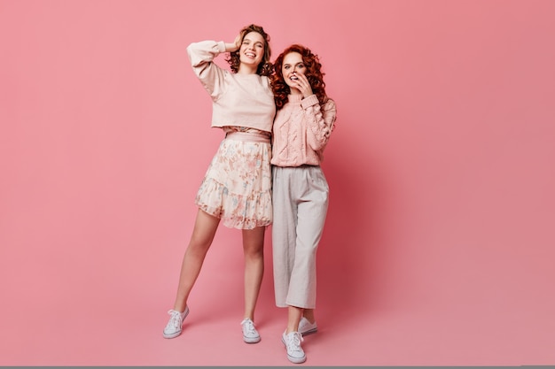 Full length view of girl in skirt posing with friend. Studio shot of two stylish young ladies standing on pink background.