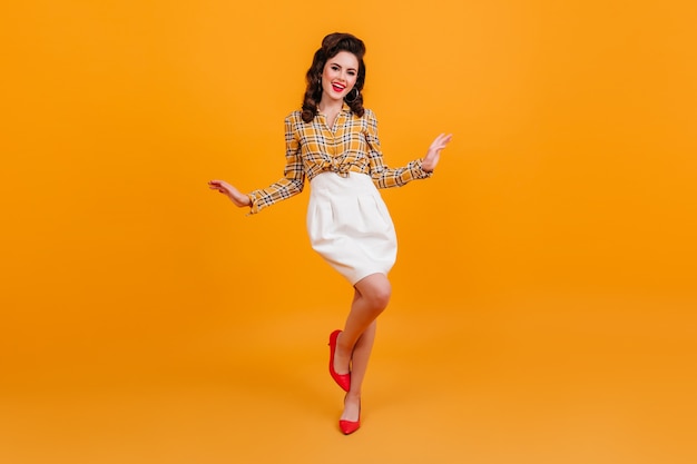 Free photo full length view of graceful curly woman in white skirt. pinup girl in checkered shirt dancing on yellow background.