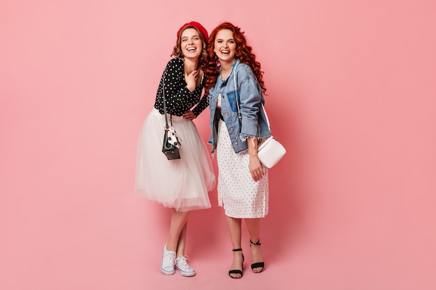 Full length view of happy sisters laughing at camera. Studio shot of trendy girls posing on pink background with smile.