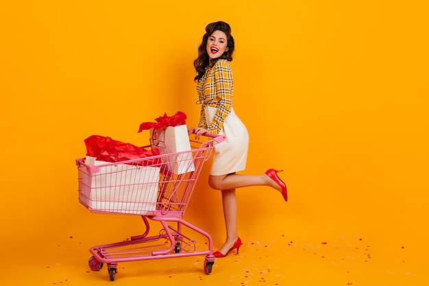 Full length view of stunning female shopper posing on yellow background. Graceful pinup girl looking at camera.