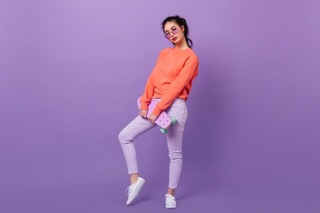 Full length view of stylish asian woman in glasses. Winsome korean model holding skateboard on purple background.