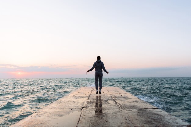 Full length view of young man in sportswear jumping with skipping rope, workout on pier