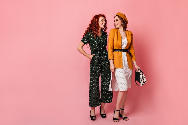 Full length view of young women in vintage outfit. Studio shot of cheerful friends talking on pink background.