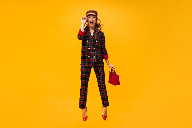 Full length woman in plaid outfit jumps on orange background and holds handbag Charming girl in eyeglasses and brown cap posing