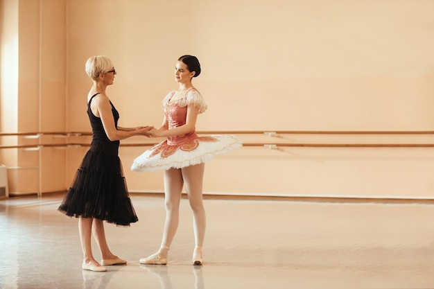 Free photo full length of young ballerina and her mature instructor holding hands while talking in ballet studio copy space