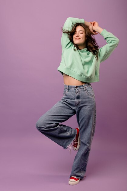 Full length young caucasian brunette girl with closed eyes in mint sweatshirt and jeans on purple studio background Concept of enjoying moment