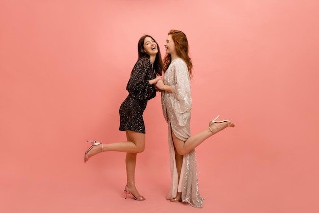 Full length young caucasian ladies in dresses stand facing each other and hold hands on pink background Lifestyle concept