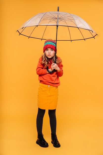 Ragazza integrale in maglione e cappello che si nascondono dietro l'ombrello e che esaminano la macchina fotografica sopra l'arancia