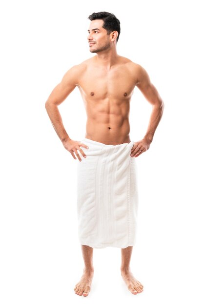 Молодой латиноамериканец в полный рост в возрасте 20 лет смотрит в сторону, стоя в полотенце на белом фоне