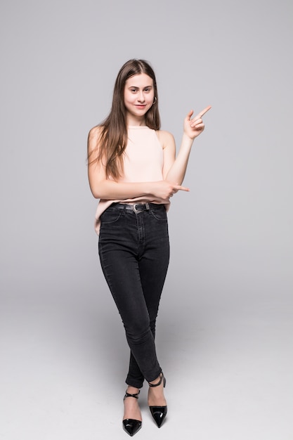Full length of young woman pointing to the side and looking there, isolated on white wall