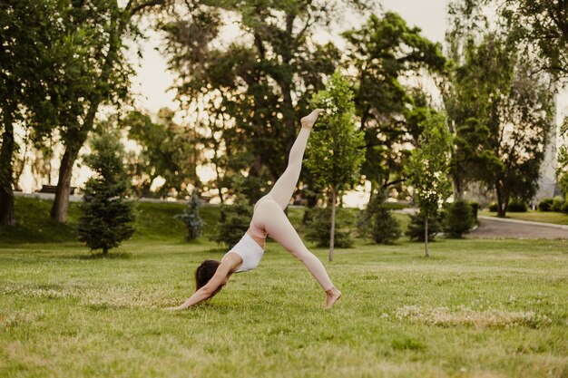 Full-length young woman practices yoga in downward-facing dog pose with one leg raised up in nature in park, concept of restoring and slowing down life.