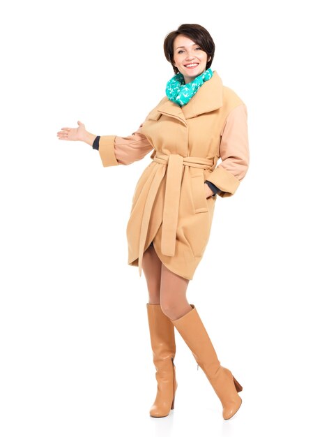 Full portrait of happy woman in beige coat with green scarf pointing on something - standing isolated on white