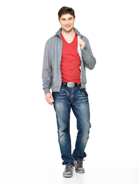 Полный портрет улыбающегося счастливого красивого человека в серой куртке, синих джинсах. Красивое положение парня изолированное на белой предпосылке.