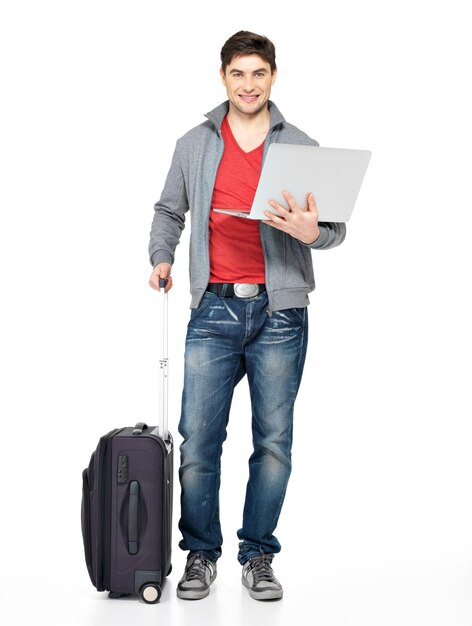Полный портрет молодого улыбающегося счастливого человека с чемоданом и ноутбуком, изолированным на белом