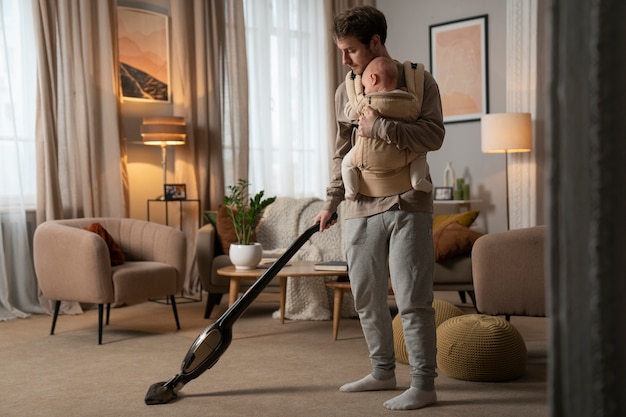 Full shot father holding baby while vacuuming