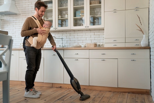 Free photo full shot father holding baby while vacuuming