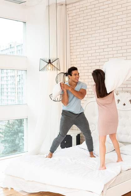 Full shot happy couple fighting with pillows in bed