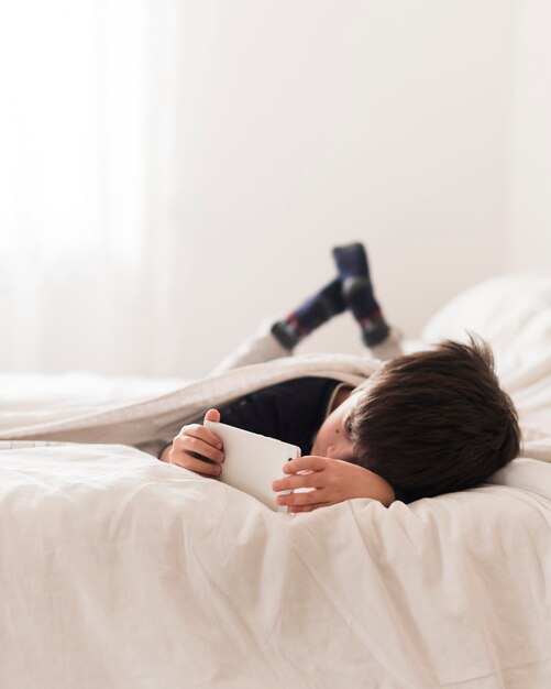 Full shot kid in bed with smartphone