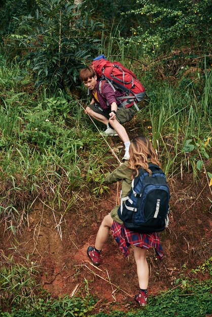 丘を登ろうとしている女性ハイカーに救いの手を差し伸べる男性ハイカーのフルショット