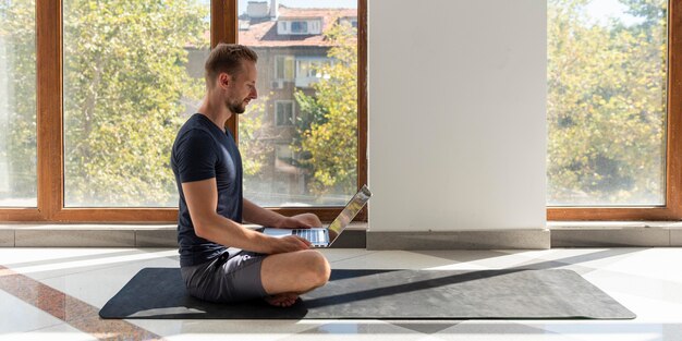 Uomo completo del colpo che si siede sulla stuoia di yoga con il computer portatile