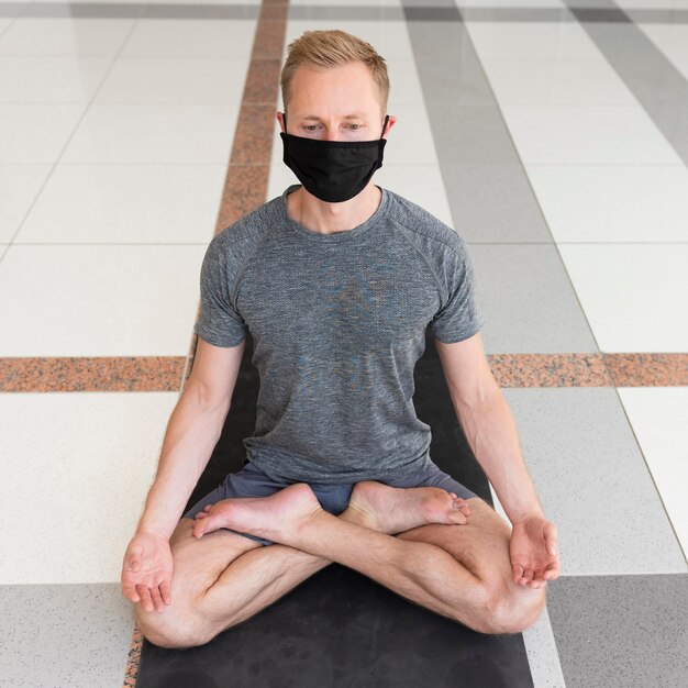 Full shot man with face mask doing sukhasana pose indoor