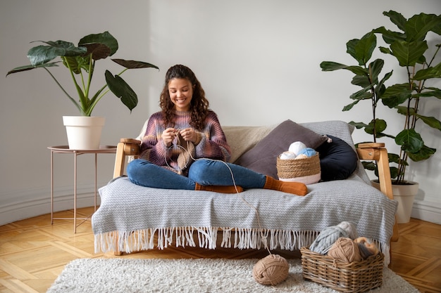 Full shot smiley young woman knitting on couch