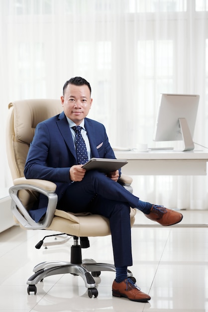 성공적인 아시아 비즈니스 임원의 전체 샷은 넓은 넓은 사무실에서 그의 고급 보스 의자에 다리를 꼬고 앉아