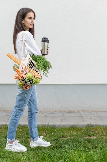 Donna della foto a figura intera che distoglie lo sguardo e che tiene borsa e termos riutilizzabili fuori