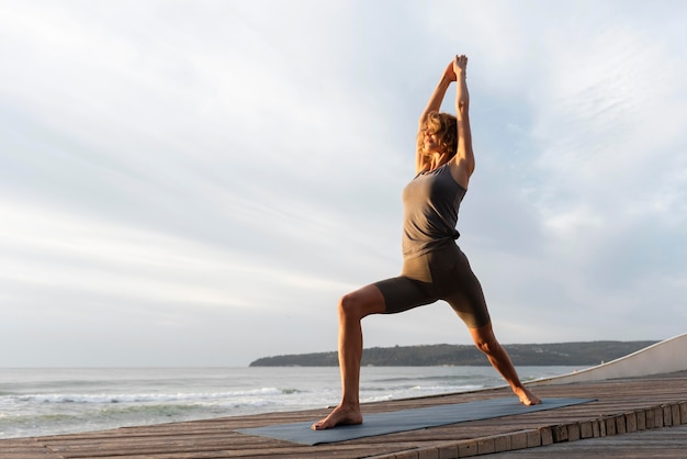 Full shot woman practicing yoga on mat near sea