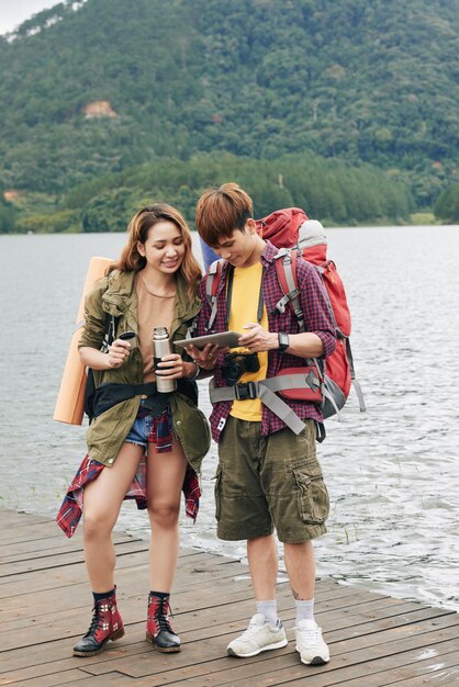 Free photo full shot of young asian couple with backpacks standing at the quay and planning their route on the tablet pc