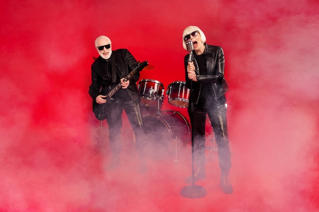 Full size photo of funky two people rock band group retired white grey hair pensioner woman vocalist sing solo man play bass guitar have fan tour isolated bright shine red color background