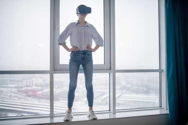 Ritratto a grandezza naturale di una donna con l'auricolare wireless per realtà virtuale in piedi sul davanzale della finestra