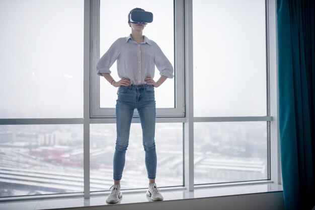 Полноразмерный портрет женщины в беспроводной гарнитуре виртуальной реальности, стоящей на подоконнике