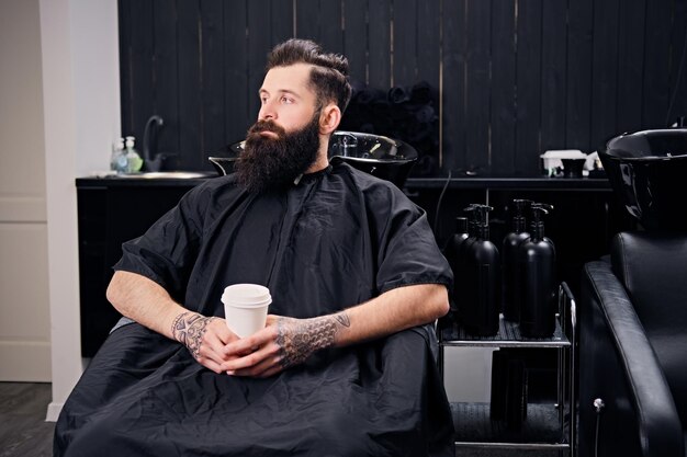 フルスロットルのひげを生やしたヒップスターの男性は、美容院でヘアスタイリングの前にコーヒーを飲みます。