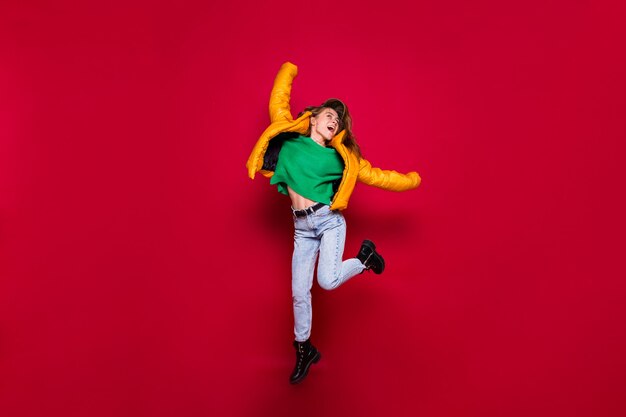 Fulllenght of happy excited girl jumping in yellow jacket and green sweater on red