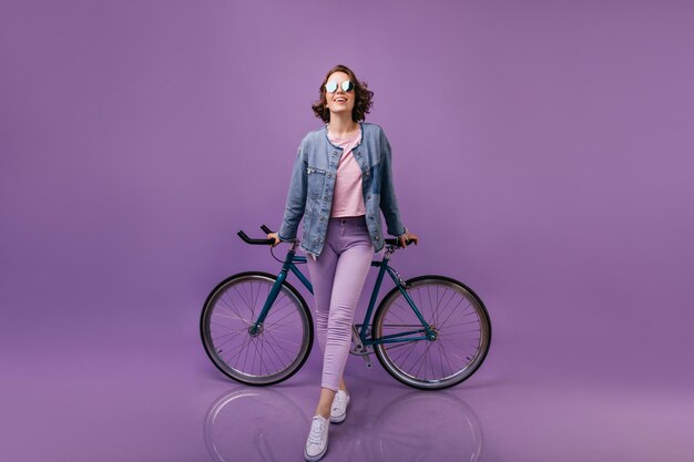 Fulllenght shot of girl in casual white shoes standing on purple background Smiling pleased woman posing near bicycle
