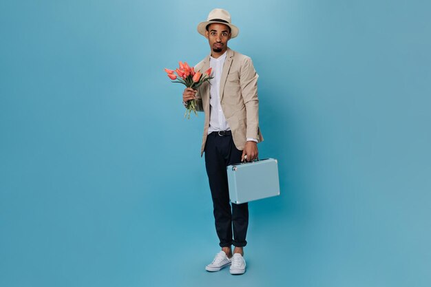 파란 배경에 꽃다발과 여행가방을 든 남자의 전신 사진 외딴 배경에서 튤립을 들고 정장을 입은 젊은 남자